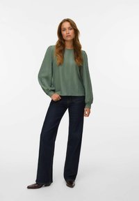 Blouse texturée vert clair à manches longues, associée à un jean évasé bleu foncé et des bottes à talons marron foncé. Design simple et décontracté.