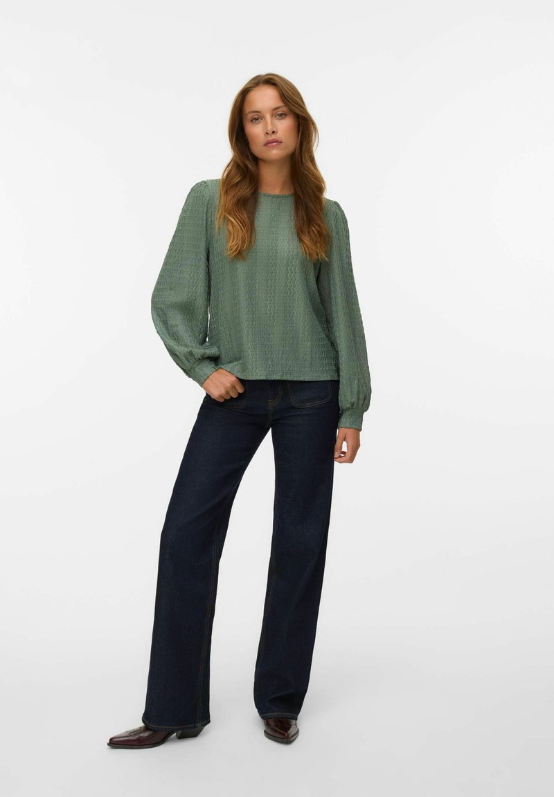 Blouse texturée vert clair à manches longues, associée à un jean évasé bleu foncé et des bottes à talons marron foncé. Design simple et décontracté.