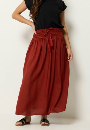 Mujer con una blusa negra y una falda de longitud hasta el tobillo en color óxido rojo con cintura ajustable con cordón y borlas, combinada con sandalias negras sin cierre.
