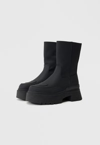 Melni platforma zābaki līdz teļiem ar masīvām rievojuma zolēm un gludu virsmas materiālu, zīmols "Replay" ārējā pusē.