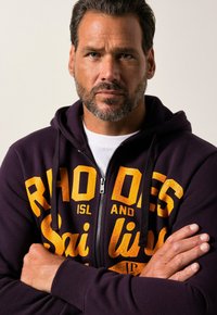 Mørk lilla zip-up hoodie med stor, sprukken gul tekst, der står "RHODES ISLAND SAILING." Har snoretræk og ribbede manchetter.