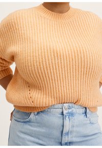 Gebreide trui in zacht perzik met geribbelde textuur, ronde halslijn en korte mouwen. Gepaarde met lichtblauwe high-waisted jeans.