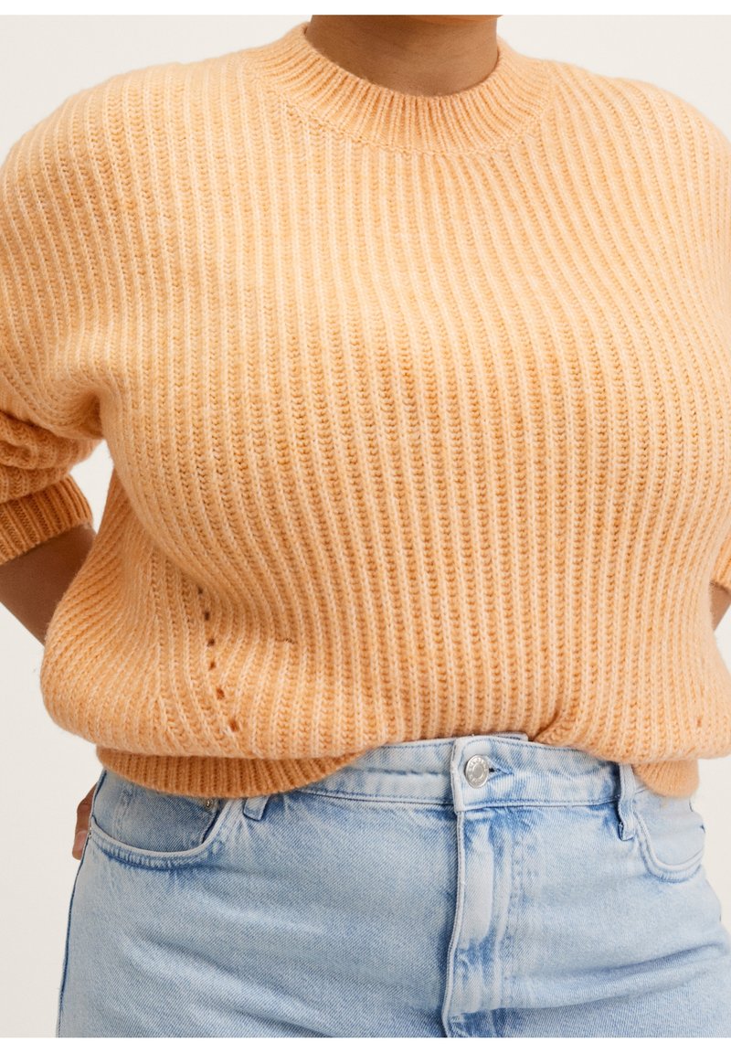 Gebreide trui in zacht perzik met geribbelde textuur, ronde halslijn en korte mouwen. Gepaarde met lichtblauwe high-waisted jeans.