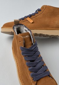 Scarpe high-top in suede marrone con lacci grigi e fodera in pelliccia, una scarpa dritta e l'altra ruotata per mostrare il motivo della suola.