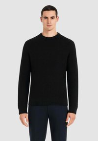 Maglione nero in maglia con scollo rotondo, polsini e orlo a costine. Presenta un tessuto dalla texture lavorata e un design aderente. Indossato sopra pantaloni scuri.