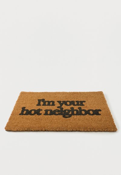 Brun kokosmåtte med sort tekst, der siger "Jeg er din varme nabo" på en ensfarvet hvid baggrund.