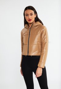Brun pufferjakke med en blank finish, lynlåslukning foran og hætte. Har horisontale syninger og et cropped design. Bæres med sorte leggings.