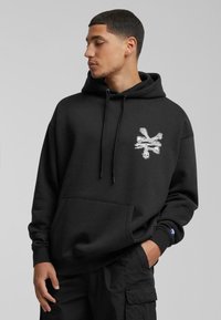 Sudadera negra con un bolsillo frontal, capucha ajustable con cordón y un logo central en blanco de una calavera y huesos. La tela parece suave y texturizada.