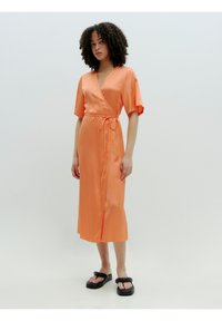 Oranje satijnen wikkeljurk met korte mouwen, strik taille en een zijsplit, gedragen met zwarte plateau sandalen. Glad textuur en subtiele glans.