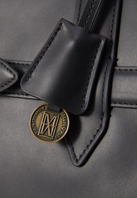 Détail d'un sac en cuir noir avec un rabat triangulaire, un médaillon logo en ton doré et des surpiqures contrastantes pour ajouter de la texture.