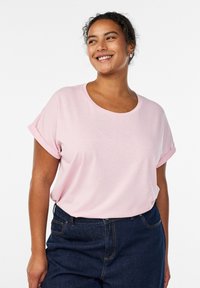 Camiseta de manga corta en color rosa claro, confeccionada en un tejido suave, con cuello redondo y mangas enrolladas, combinada con pantalones vaqueros de talle alto en color oscuro.