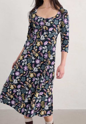 Femme portant une robe trapèze fleurie bleu marine avec des manches trois-quarts et un col carré sur un fond clair uni.