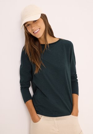 Chemise à rayures, vert foncé, à manches longues en tissu doux, assortie à une casquette beige claire et un short de couleur claire, les mains dans les poches.