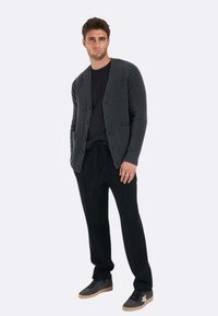 Cardigan grigio a maglia con chiusura a bottoni e due tasche frontali, indossato sopra una maglietta nera, abbinato a pantaloni neri e sneakers grigie.