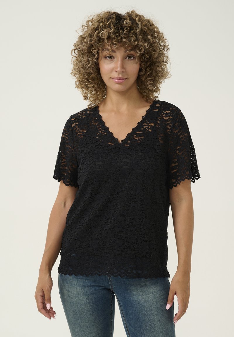 Black lace top with short sleeves and V-neckline. Scalloped edges and floral pattern create a delicate texture. Worn with blue jeans.