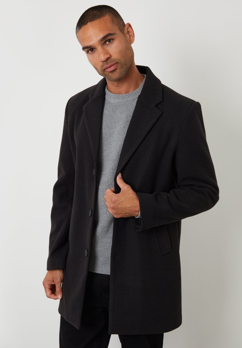 Sort uld-blandet frakke med klapkrave, en- eller to- knaps design, båret over en grå ribbet sweater. Glat tekstur.