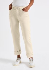 Pantalones de tiro alto color beige claro con puños enrollados, dos bolsillos frontales y cierre de botón. Hechos de mezclilla con una textura suave. Combinados con zapatillas blancas.