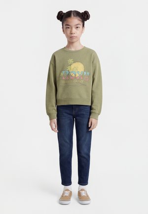 Djevojka s dva svemirska pundža nosi maslinasto zelenu majicu dugih rukava s natpisom "Roxy sunsets chaser", tamne traperice, bijele čarape i svijetlosmeđe tenisice, stoji mirno.