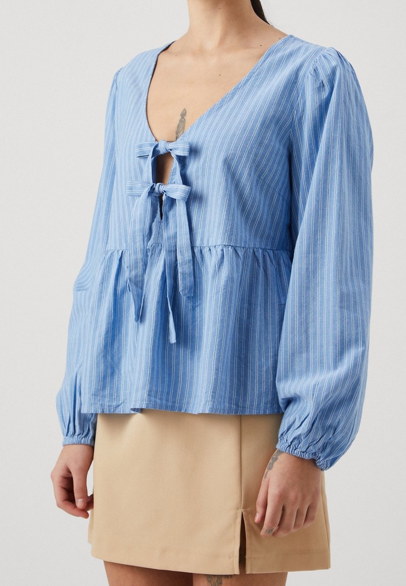 Ljusblå, randig blus med V-ringning och frontknut. Har långa, puffiga ärmar och inhämtad midja, tillverkad av ett mjukt tyg.