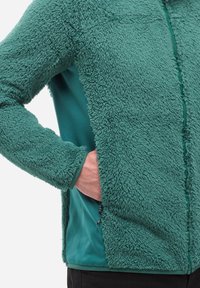 Türkisgrüne, flauschige Jacke mit glattem Stoffeinsatz an der Seite, Frontreißverschluss und Händen in den Seitentaschen vor einem weißen Hintergrund.