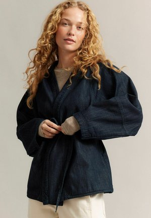 Kobieta z kręconymi blond włosami zawiązuje pasek na ciemnej dżinsowej kurtce na tle beżowego topu i białych spodni na neutralnym tle.