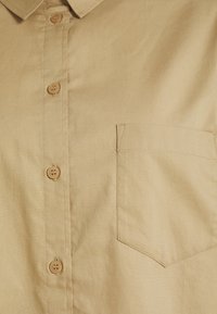 Chemise beige boutonnée en tissu doux, dotée d'un col classique, de boutons marron et d'une poche poitrine gauche unique avec un ourlet droit.