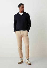 Polo a maniche lunghe blu navy, pantaloni della tuta beige con cintura elastica, patch con logo e sneakers bianche con dettagli neri e suola testurizzata.