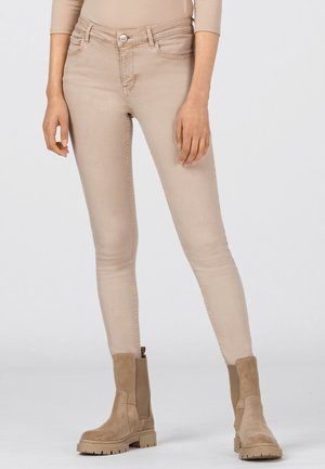 Vrouw draagt beige skinny broek en bijpassende beige suède enkellaarsjes, staand tegen een effen lichte achtergrond.