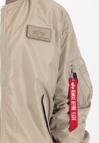 Bež nailonist bomber-jakk, millel on rinnal plekk, varrukal lukuga tasku ja punane "Eemalda enne lendu" silt.