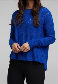Blauer Strickpullover mit strukturiertem Blattmuster, langen Ärmeln und leicht lockerem Schnitt, kombiniert mit schwarzen, eng anliegenden Hosen.