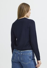 Pull en maille bleu marine à manches longues et col rond. Texture lisse, silhouette ajustée et détails minimaux. Porté avec un jean taille haute.