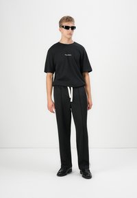 Black short-sleeve T-shirt with subtle logo, paired with black trousers featuring a drawstring waist. Model wears black sunglasses and shoes.