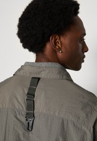 Gray lightweight jacket with a textured finish, featuring a strap and clip detail at the back, solid collar, and subtle stitching.