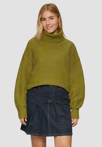 Olijfgroen, oversized gebreide trui met hoge kraag, gecombineerd met een donkere spijkerrok met plooien en zichtbare stiksels.