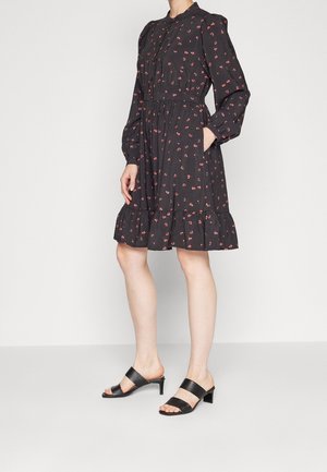 Femme portant une robe noire à manches longues à motif floral avec un ourlet à volants et des sandales à talons noires, debout devant un fond uni.