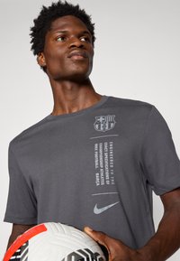 T-shirt gris à manches courtes avec col rond, arborant un écusson de Barcelone et le logo Nike, tissu texturé et design imprimé sur le côté.