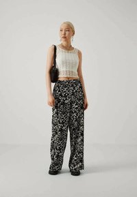 Woman with short blond hair wears white lace sleeveless top, black-and-white patterned wide-leg pants, black shoes, and a black shoulder bag.