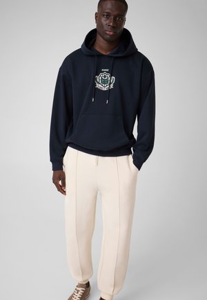 Mann, der einen dunkelblauen Hoodie mit einem grün-weißen Emblem, cremefarbene Jogginghosen mit vorderen Nähten und beige Sneaker trägt, steht vor einem grauen Hintergrund.