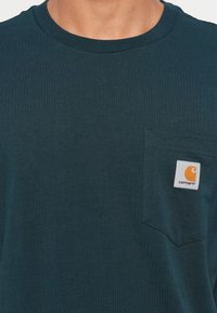 Donkerteal katoenen T-shirt met een ronde hals, voorzien van een borstzak en een vierkant wit label met een oranje logo.