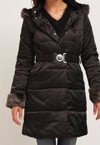 Černá puffa bunda s kapucí lemovanou umělou kožešinou, páskem v pase, zipem, diamantovým prošíváním a dvěma předními kapsami.