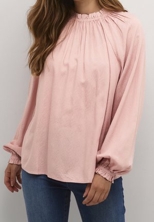 Vrouw draagt een lichtroze blouse met lange mouwen, een geplooide halslijn en manchetten, gecombineerd met een blauwe spijkerbroek, staand tegen een effen lichte achtergrond.