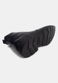 Botín de cuero negro hasta el tobillo con un upper liso, suela de goma texturizada y un tacón bajo. Cuenta con un tirador en la parte posterior para facilitar su puesta.