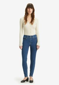 Cárdigan crema con canalé y cierre de botón, y jeans skinny azul de tiro alto, combinados con zapatos planos negros. Diseño simple y ajustado.