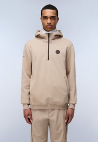 Sudadera con capucha beige de media cremallera, con parche de logo en el pecho y puños acanalados, combinada con pantalones beige a juego. Textura suave.