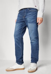 Jeans in denim blu leggero con un design classico a gamba dritta, abbinati a sneaker bianche e a un maglione grigio con texture.