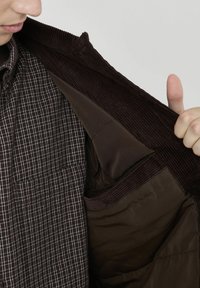 Veste en velours côtelé marron avec une doublure intérieure lisse marron, portant une chemise à carreaux marron et blanc en dessous, montrant une poche de rangement.