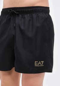 Μαύρα μαγιό σορτς από απαλό ύφασμα, με ελαστική μέση και κορδόνι, και με χρυσό λογότυπο στο τελείωμα.