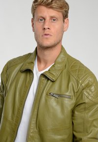 Olivgrüne Lederjacke mit gesteppten Schulterakzenten, Reißverschlusstaschen und Stehkragen. Getragen über einem weißen T-Shirt.