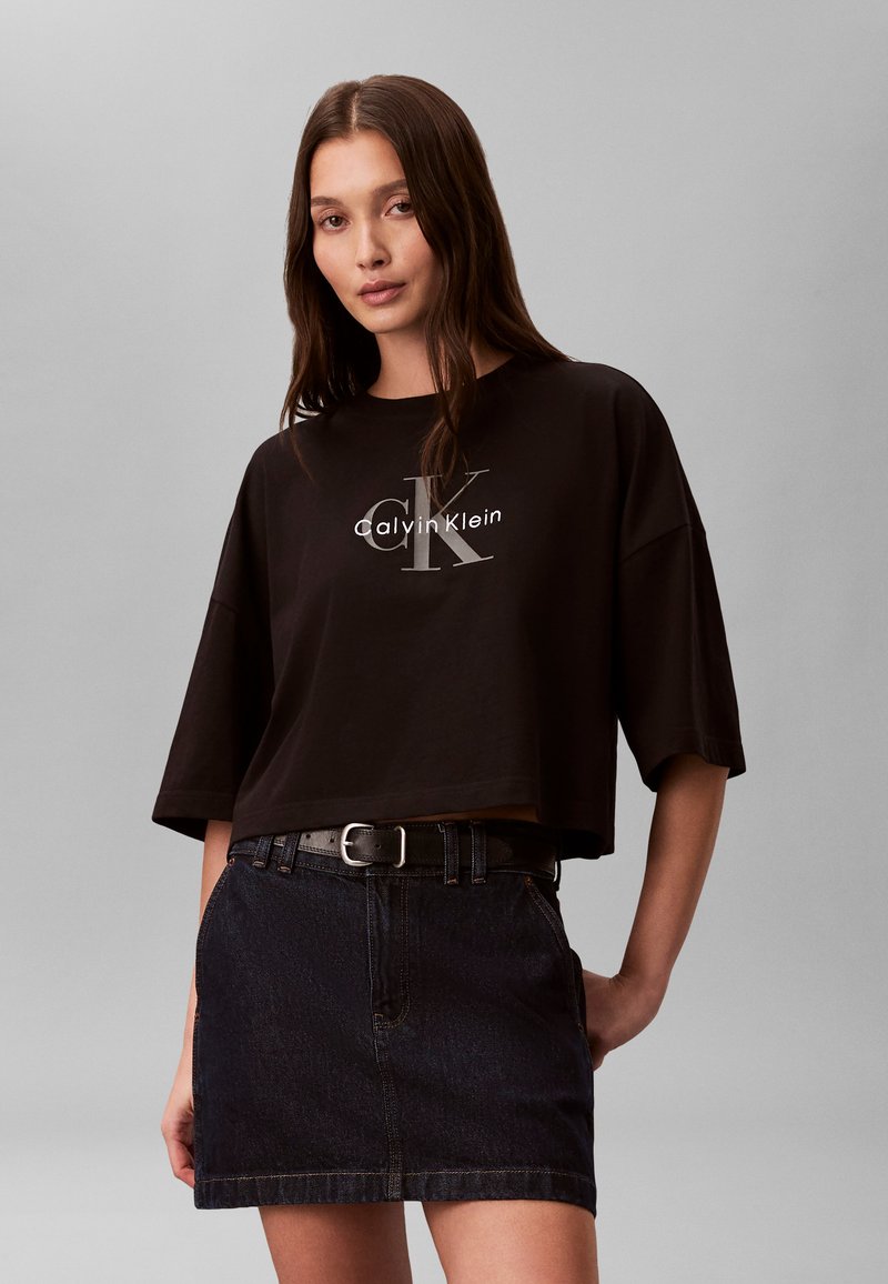 Γυναίκα που φοράει μαύρο κομμένο T-shirt Calvin Klein και σκουρόχρωμη φούστα από τζιν με μαύρη ζώνη, στέκεται μπροστά σε ουδέτερο γκρίζο φόντο.