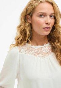Weiße Bluse mit einer Spitzenblende, die florale Muster zeigt, runder Ausschnitt und fließendem Stoff. Weiche, leichte Textur, lange Ärmel.